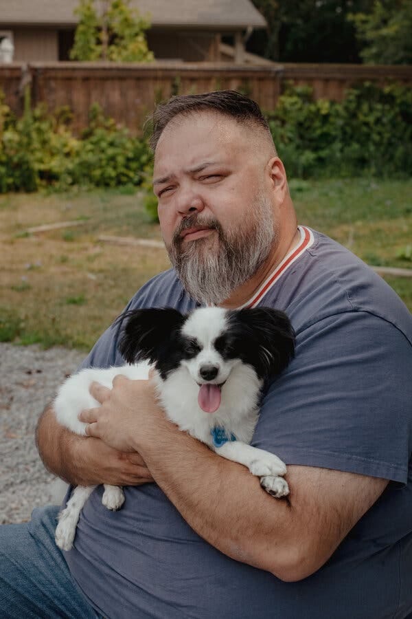 Allan Brooks, wearing a blue shirt with red and white stripes around the neck, holds a black and white dog in his arms. Behind him is a grassy yard with a wooden fence.
