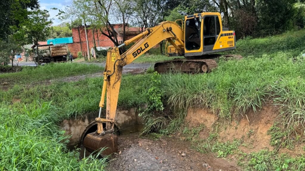A imagem apresenta uma escavadeira amarela SDLG realizando trabalho de limpeza ou manutenção em um canal de drenagem. O equipamento está sobre um terreno gramado, com o braço estendido até o fundo do canal, enquanto ao fundo há construções simples e vegetação. Essa cena representa uma ação de obras de drenagem urbana ou manejo de águas pluviais. A imagem apresenta uma escavadeira amarela SDLG realizando trabalho de limpeza ou manutenção em um canal de drenagem. O equipamento está sobre um terreno gramado, com o braço estendido até o fundo do canal, enquanto ao fundo há construções simples e vegetação. Essa cena representa uma ação de obras de drenagem urbana ou manejo de águas pluviais.