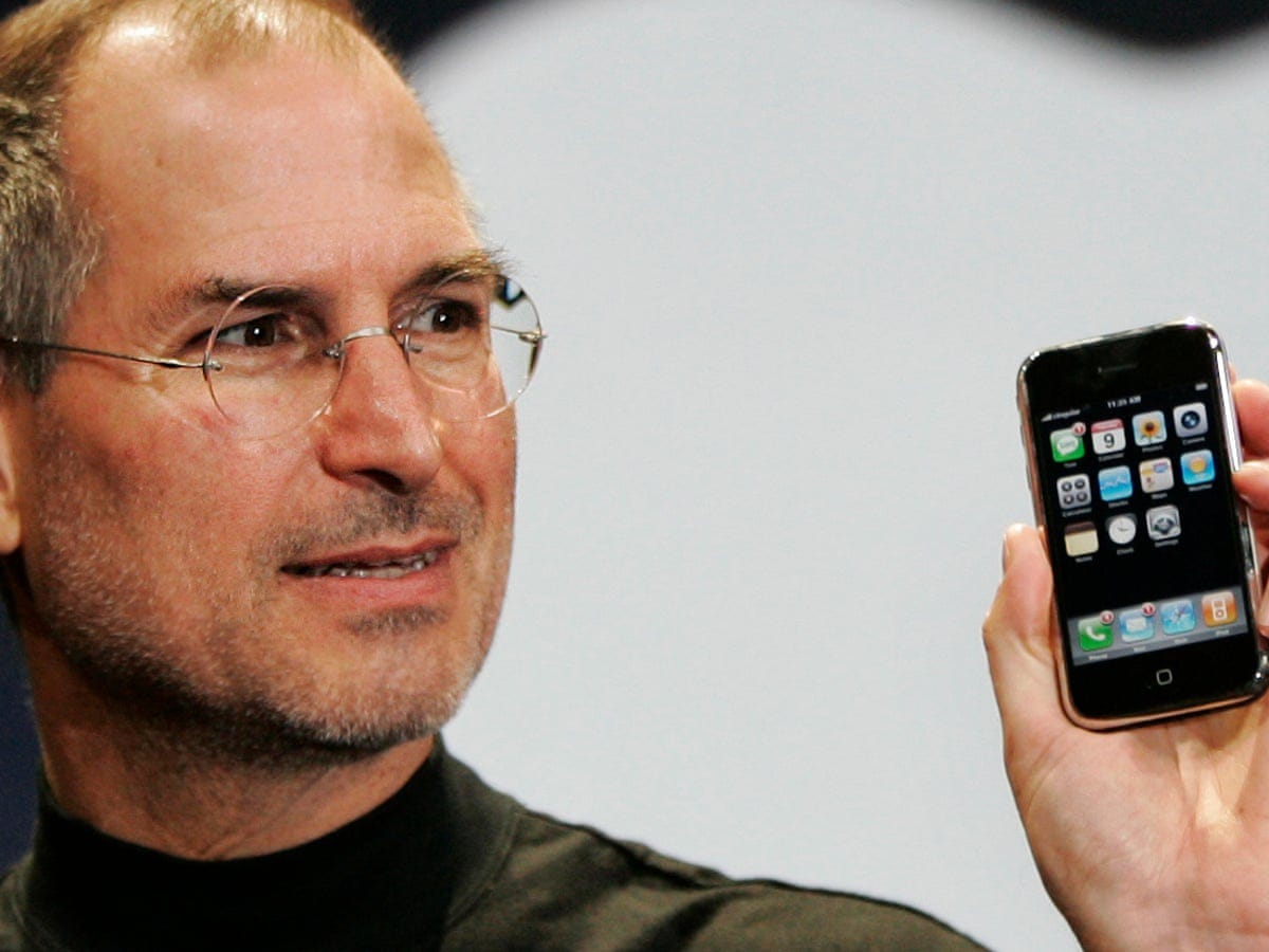 Steve Jobs presents the first iPhone to an audience, holding the device upright as if introducing a revolutionary piece of technology.