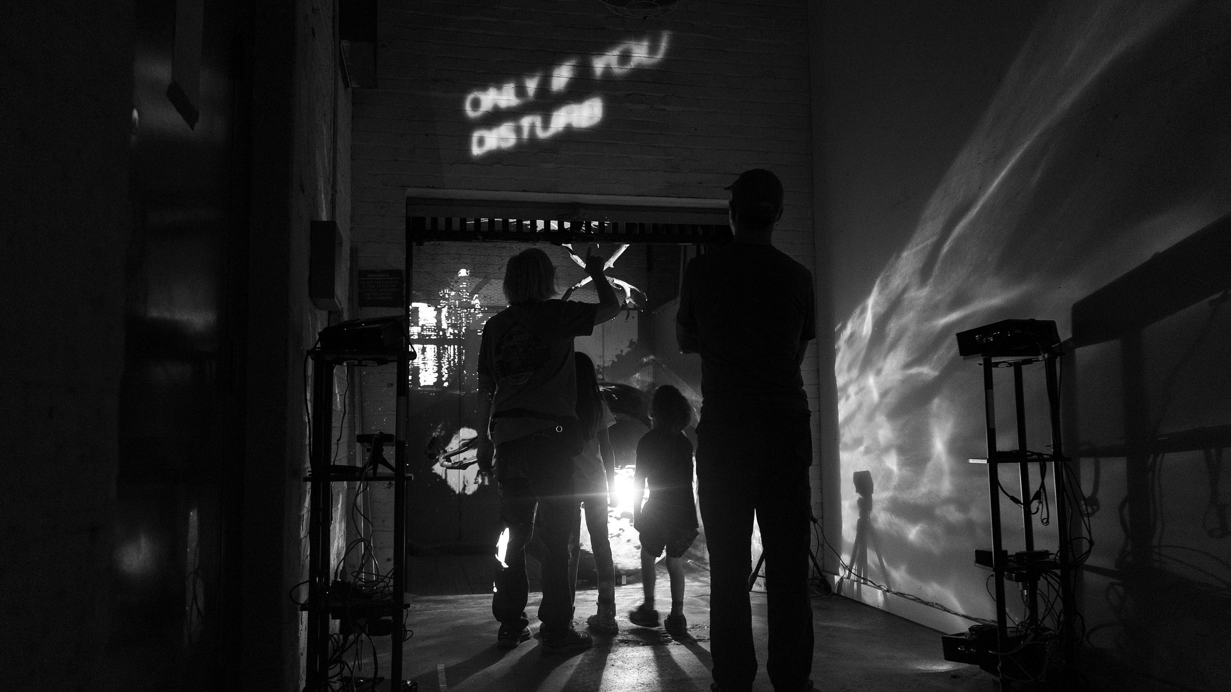 This black and white image shows two adults and two children from behind. They are standing in front of the art installation. The brightness of the projectors renders them into silhouettes.