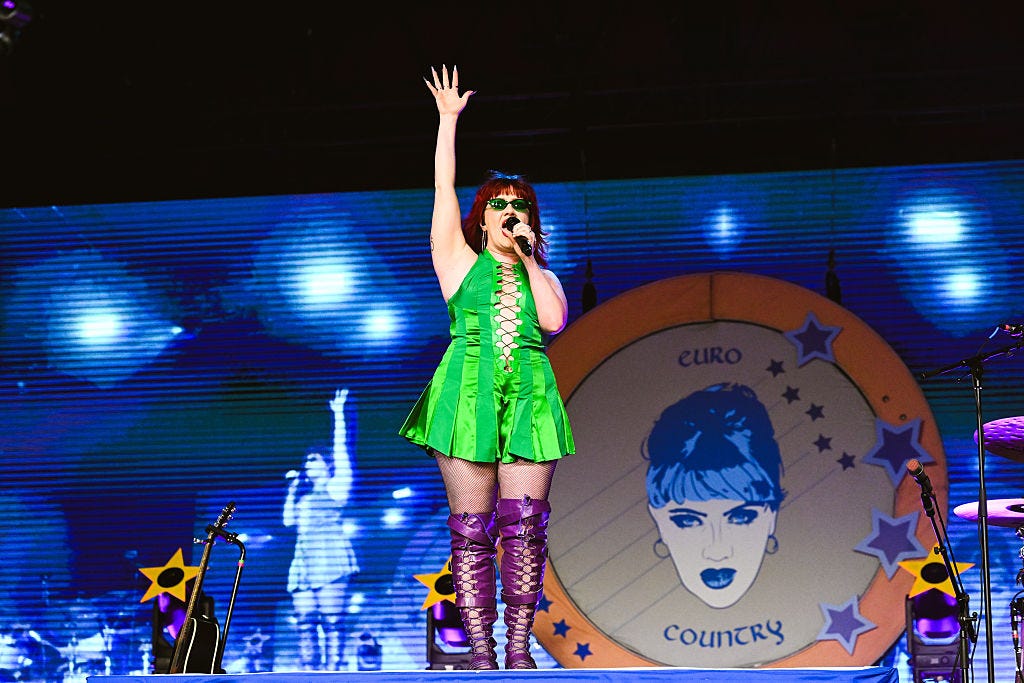 A performer in a green dress and purple boots sings on stage with one arm raised, in front of a large backdrop featuring a stylized face and the words "Euro Country.
