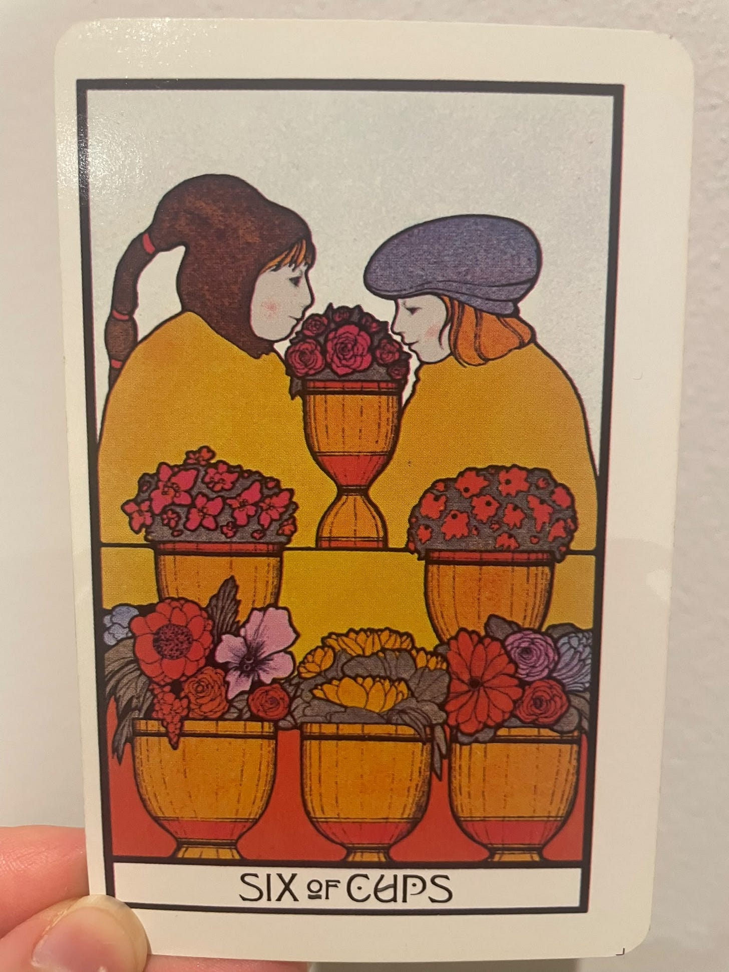 The Six of Cups tarot card, illustrated in a vintage-inspired style. Two young figures dressed in yellow face each other across a golden cup filled with red roses. Five additional golden cups overflow with colourful flowers in the foreground, symbolising nostalgia and childhood memories. The Six of Cups tarot card, illustrated in a vintage-inspired style. Two young figures dressed in yellow face each other across a golden cup filled with red roses. Five additional golden cups overflow with colourful flowers in the foreground, symbolising nostalgia and childhood memories.