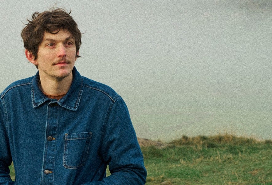 A grainy photo of Chris Brain. He is sitting in the countryside on a grey day, wearing a denim overshirt that is buttoned up to the top.