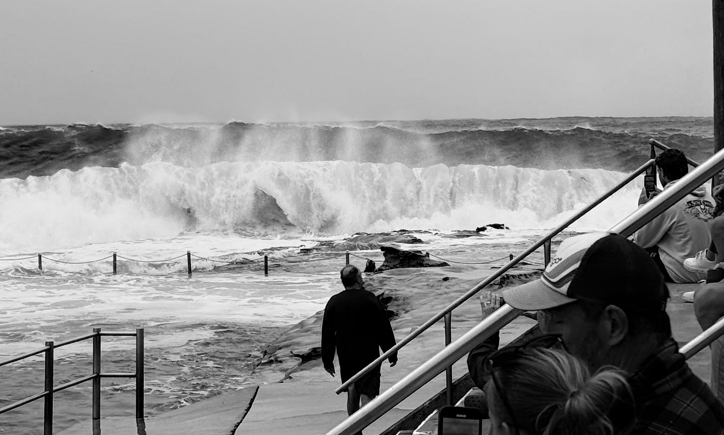 Black and white image of a set of large waves breaking on the headland rocks and about to engulf the ocean pool with a man walking toward the break and people in the foreground sitting on the steps and watching the surf..