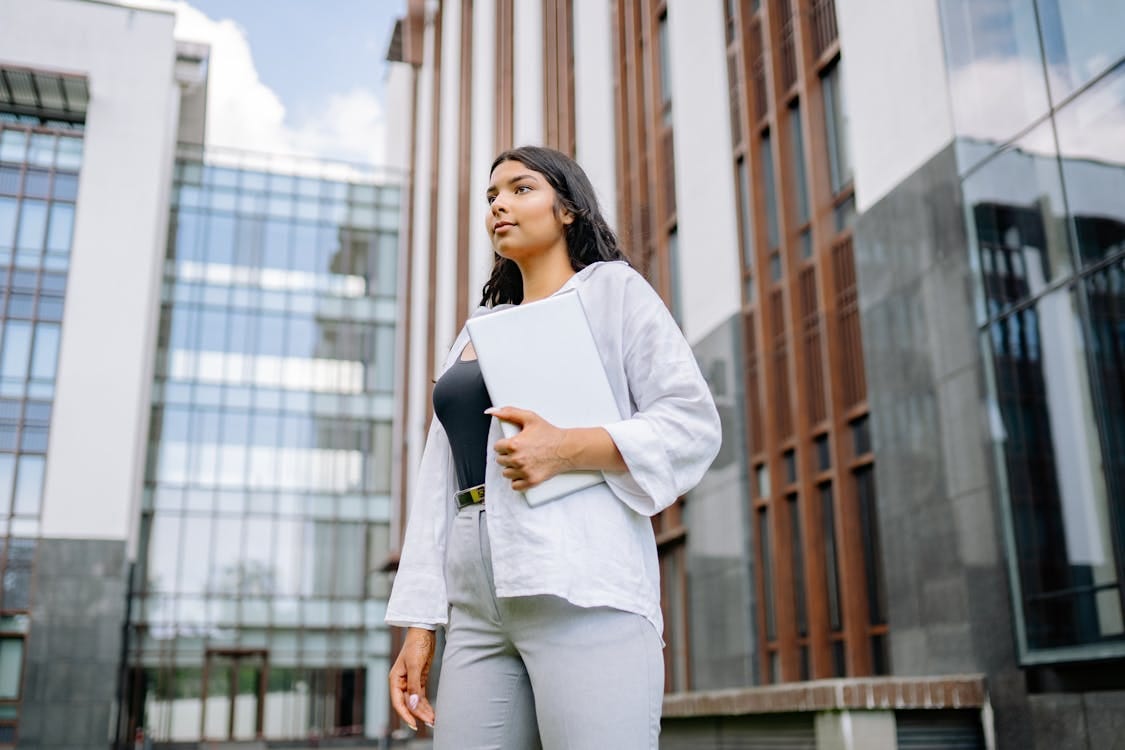 Free Confident businesswoman stands holding a laptop in front of a modern building. Stock Photo