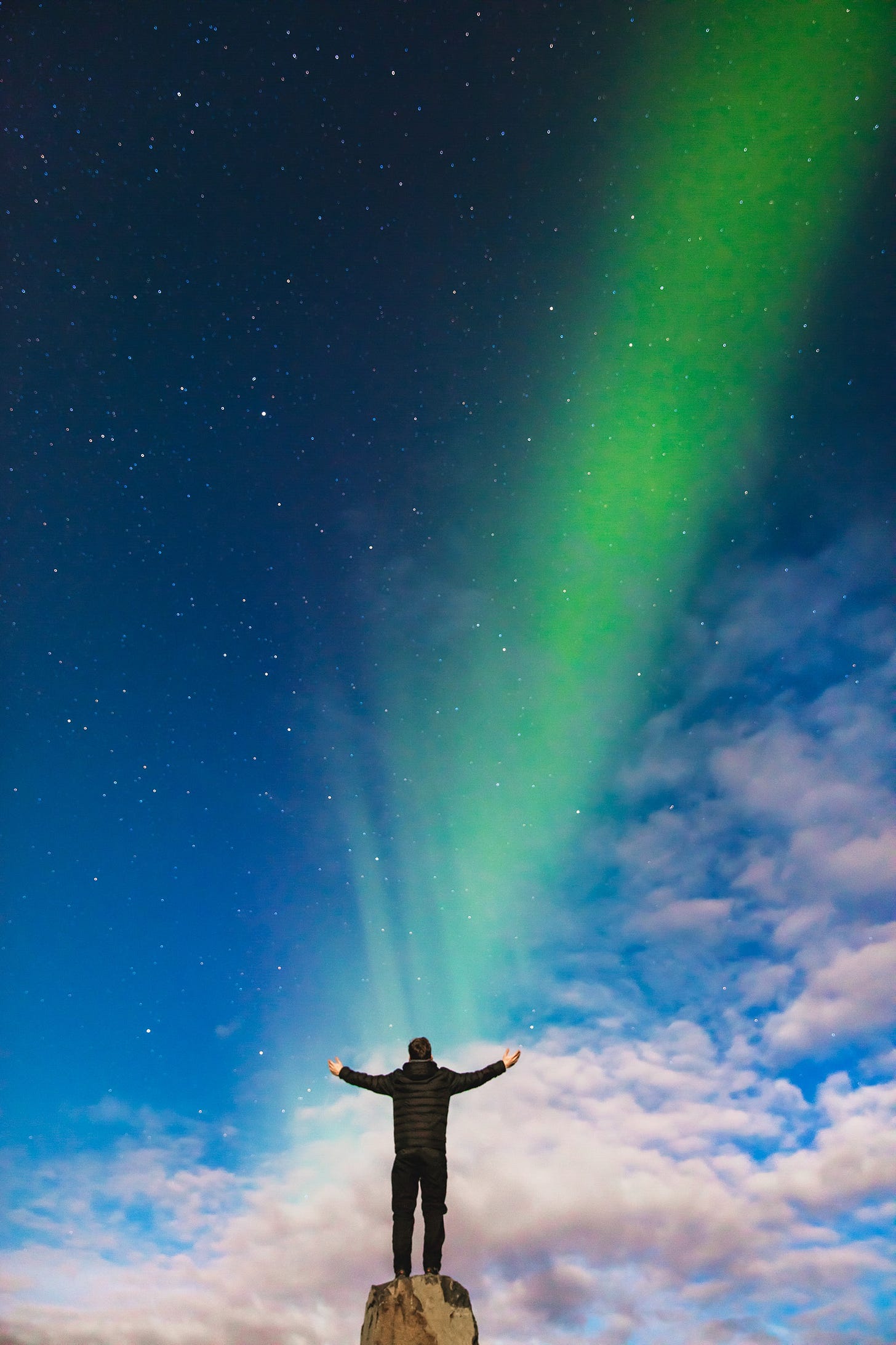 Color photograph of a man wearing a black parka, his back to the camera, facing a darkening sky filled with pink clouds, stars, and green aurora Color photograph of a man wearing a black parka, his back to the camera, facing a darkening sky filled with pink clouds, stars, and green aurora