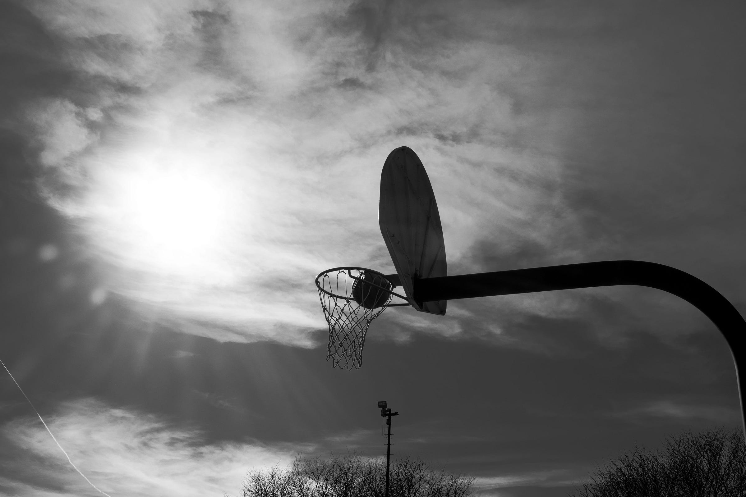 A black-and-white photograph of a basketball hoop silhouetted against a dramatic sky, with the sun shining through scattered clouds. A basketball is caught in the net, frozen in motion. The curved arm of the hoop extends into the frame, creating a striking contrast against the soft, diffused light. Bare tree branches and a tall floodlight pole appear in the distance, adding depth and scale to the scene. The image evokes a sense of nostalgia, resilience, and the timeless nature of the game.