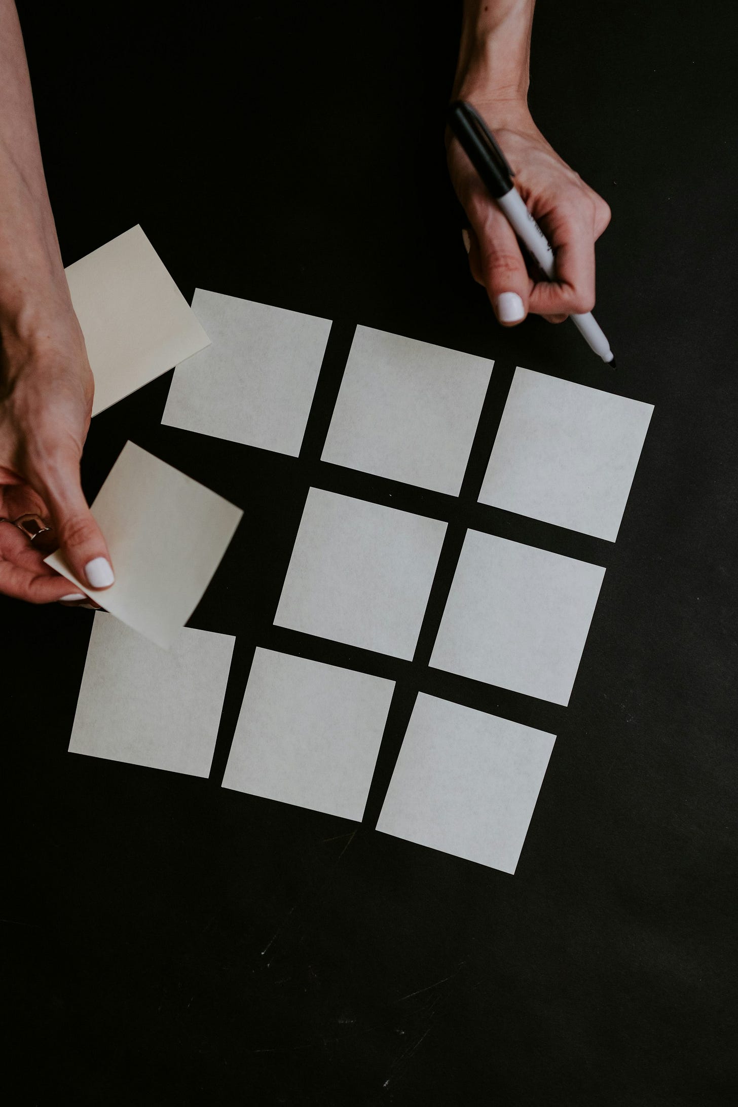 White square post-it notes rest on a black surface, three across, two below, and three beneath that. A woman's hand moves to add a third to the middle row, and a small pile of additional square post-its rest beneath her wrist to the left. White square post-it notes rest on a black surface, three across, two below, and three beneath that. A woman's hand moves to add a third to the middle row, and a small pile of additional square post-its rest beneath her wrist to the left.
