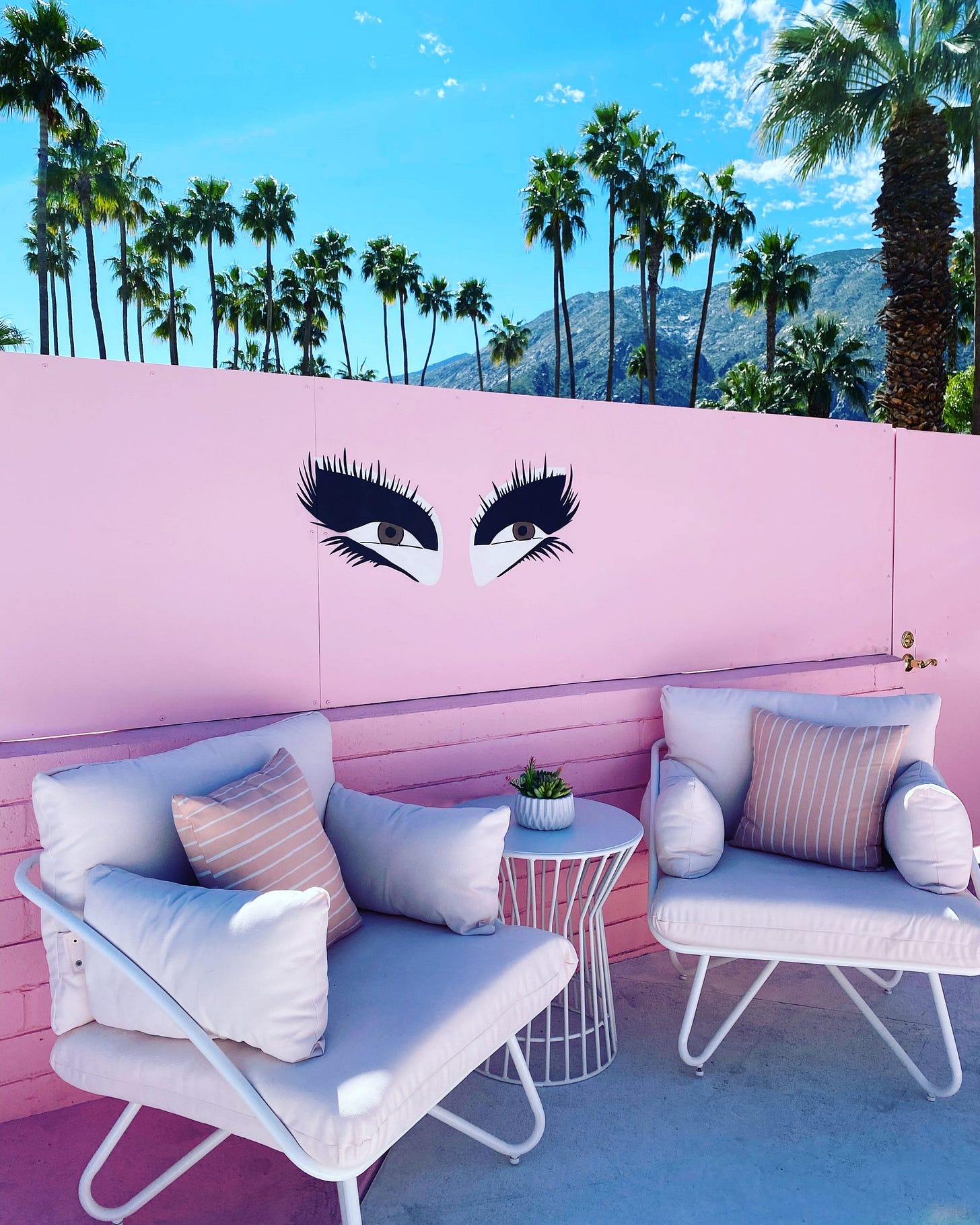 Outdoor seating area with two white cushioned chairs and a small table on a sunny patio. Each chair has a purple patterned pillow on them. Light pink wall adorned with painted eyes that resemble Trixie Mattel's makeup. Palm trees and mountains in the background. Outdoor seating area with two white cushioned chairs and a small table on a sunny patio. Each chair has a purple patterned pillow on them. Light pink wall adorned with painted eyes that resemble Trixie Mattel's makeup. Palm trees and mountains in the background.