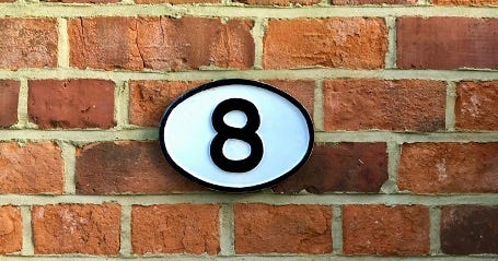 Photo of a brick wall. There is a white plaque on the wall with a black number eight on it. 