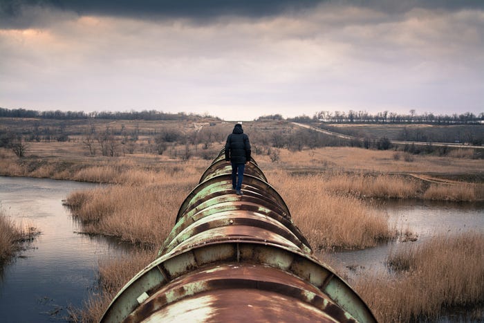 A jacketed man walks down the length of a rusty old pipeline. A jacketed man walks down the length of a rusty old pipeline.