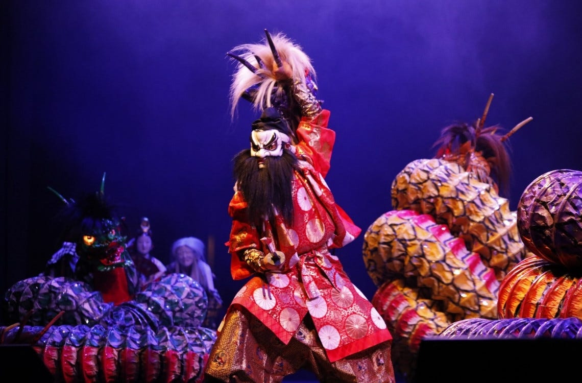 A performer in a vibrant red costume and fierce mask raises a sword during a traditional Japanese kagura dance, with elaborate dragon props surrounding the stage.