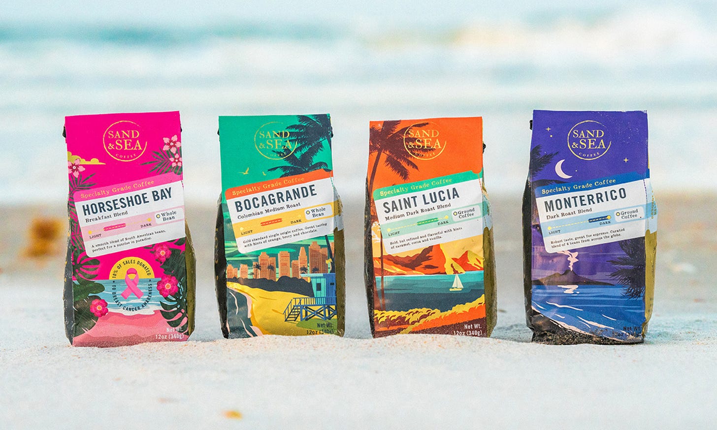 Four colorfully decorated coffee bags rest on the sand with the surf blurred in the background. The bags from left are predominantly pink, green, orange, and purple. Four colorfully decorated coffee bags rest on the sand with the surf blurred in the background. The bags from left are predominantly pink, green, orange, and purple.