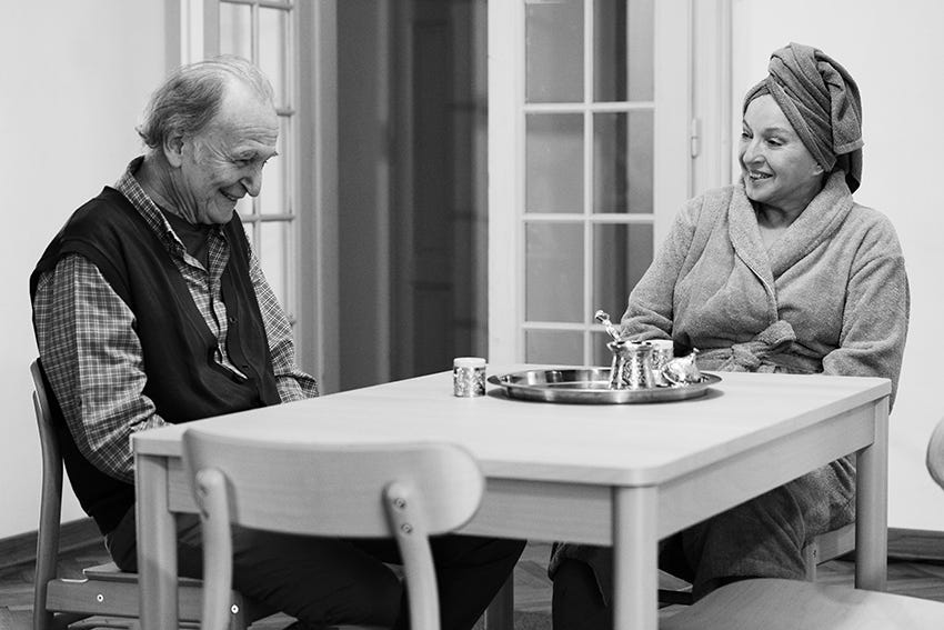 A scene from They Have All Gone. Sadika and Azem sit at a table. Her hair is wrapped in a towel and she is wearing a bathrobe. They are both laughing.