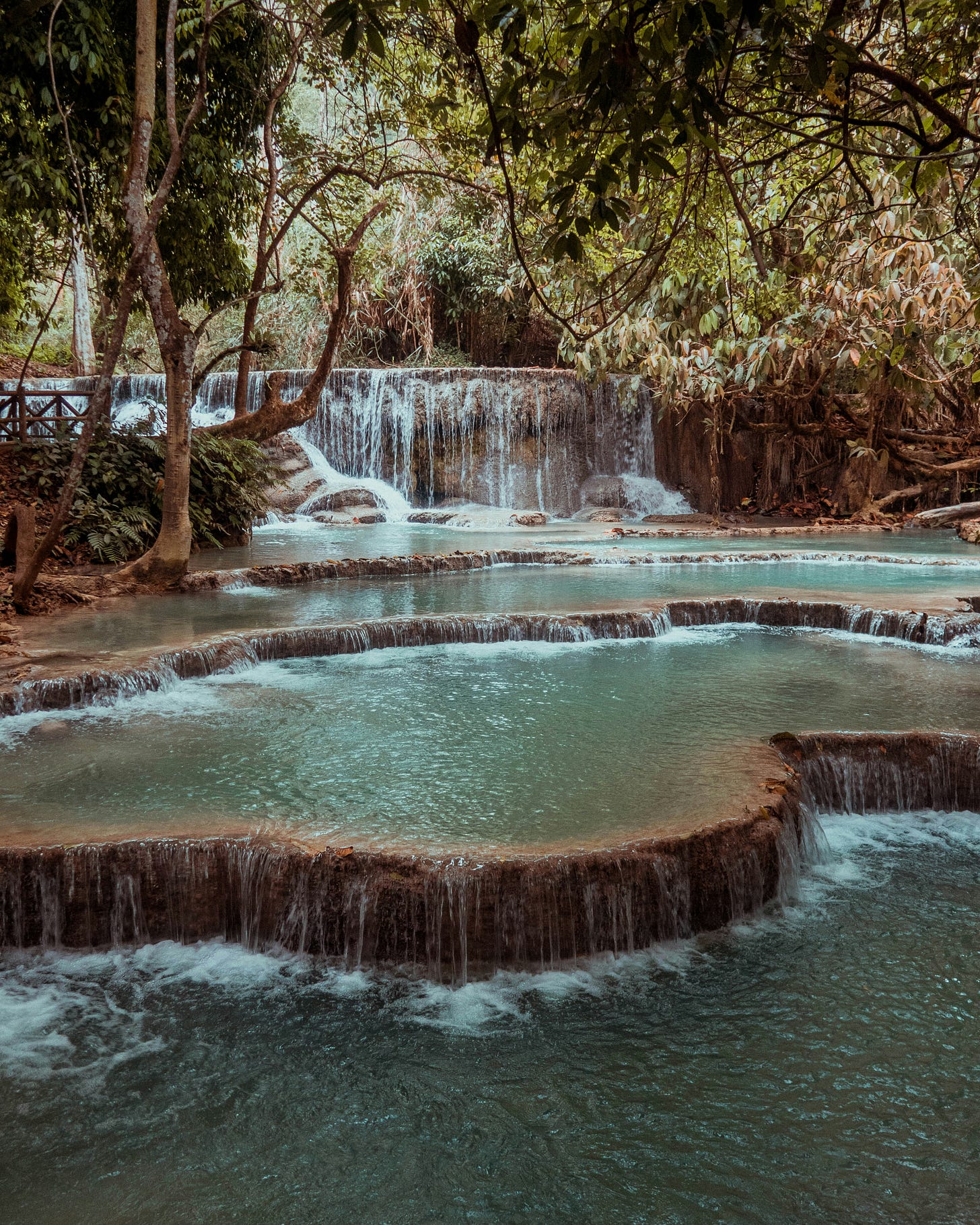 A multi-tiered waterfall cascading down limestone formations into a bright turquoise pool in a dense jungle.