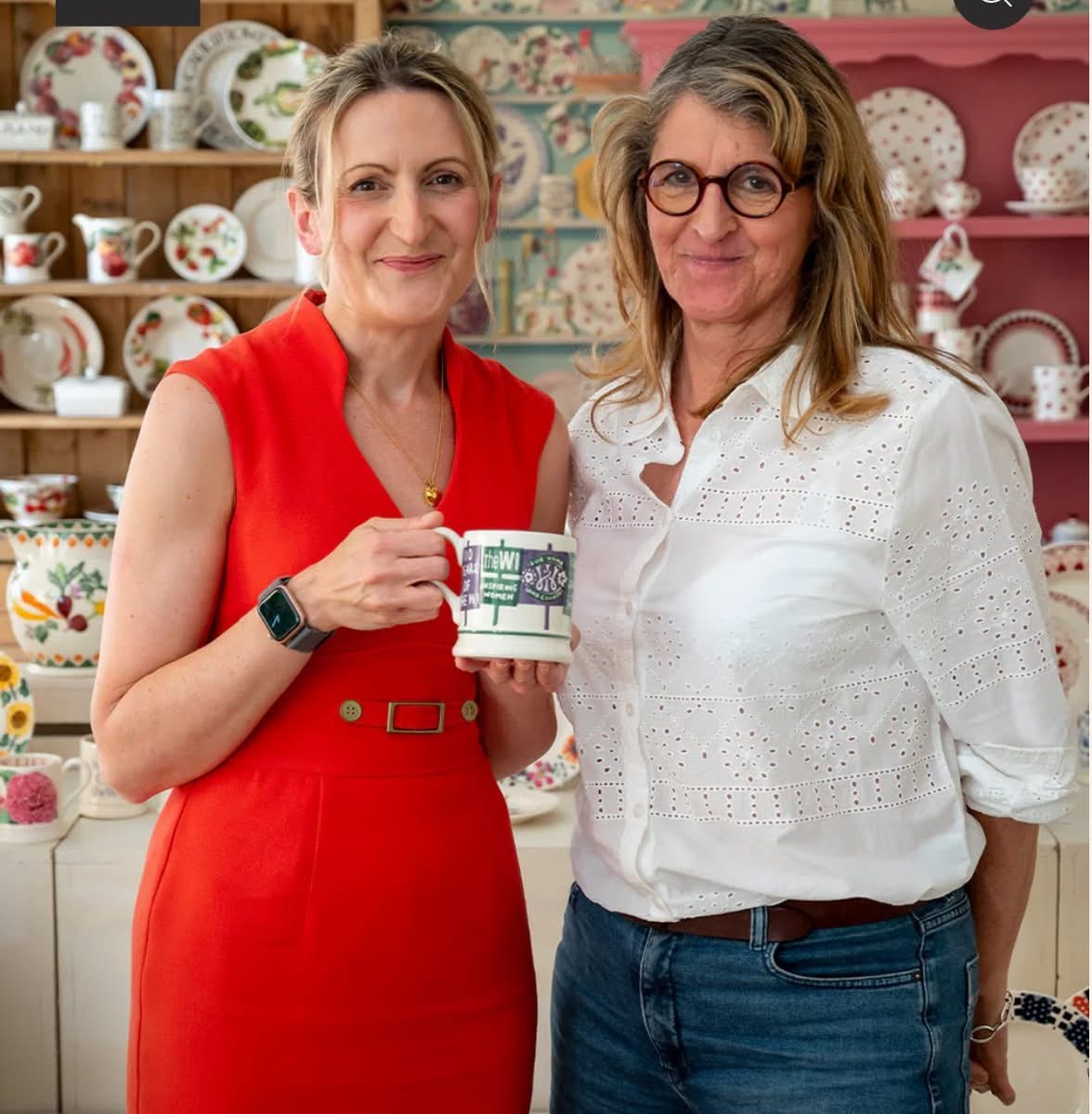 Two women standing in a pottery workshop.  Melissa Green is on the left, blonde slim woman with bright red sleeveless dress.  She is holding the new WI mug created by Emma Bridgewater.  Emma is standing next to her with a white blouse and she is wearing glasses.  The dressers behind them are full of various pottery designs including fruit, spots etc