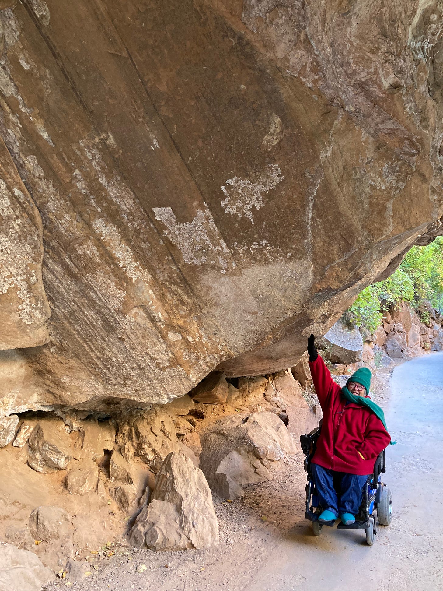 A white woman using a power chair bundled up in warm clothes underneath a rock overhang and holding her hand up against it as if she was supporting the weight. 