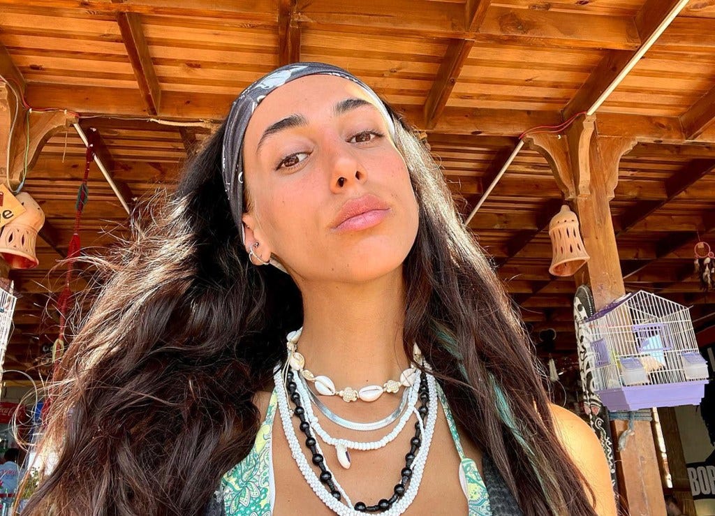 Tali Biner, an Oct. 7 survivor, wearing a patterned bandana, layered necklaces, and a printed top, looking directly at the camera.