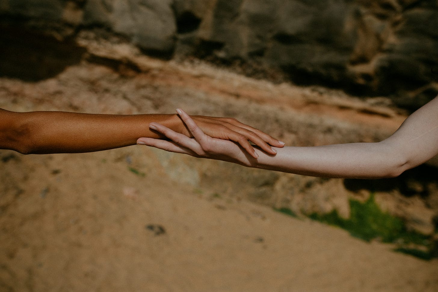 Two arms reach across the frame, hands lightly touching, over sand and rock.