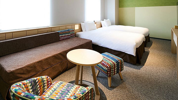 The modern, bright reception area of Hotel Intergate Kanazawa featuring a colorful geometric tiled wall and a long minimalist wooden front desk. An accessible hotel bathroom vanity area with a wide white countertop, a square vessel sink, and open wooden shelving containing towels and a small refrigerator. A spacious accessible hotel room featuring two twin beds, a large brown sofa, and a small round table with colorful patterned chairs and stools. An accessible bathroom featuring a bathtub with a handheld shower head, a sink, and a toilet with grab bars against light wood-textured walls.