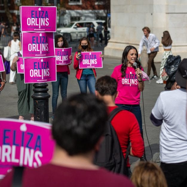 Bright pink "Eliza Orlins" signs adown a light pole as the candiate addresses a crowd next to it as others hold pink signs too