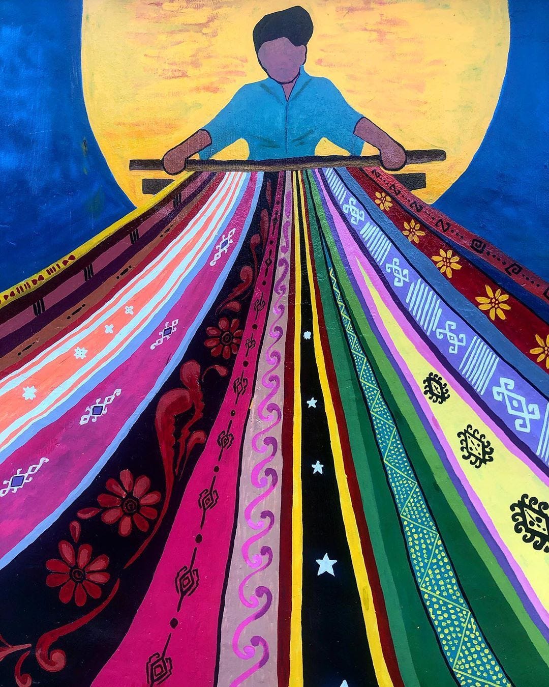 A vibrant painting depicting a stylized, faceless figure in a light blue shirt holding a horizontal wooden beam. Behind the figure's upper body is a massive, glowing golden sun filling the center against a deep blue sky. Spreading downwards from the beam like a wide fan or apron are dozens of long, tightly arranged strips of fabric, each featuring unique, intricate folk patterns and bright colors including pink, red, black with white stars, green, yellow, and purple.