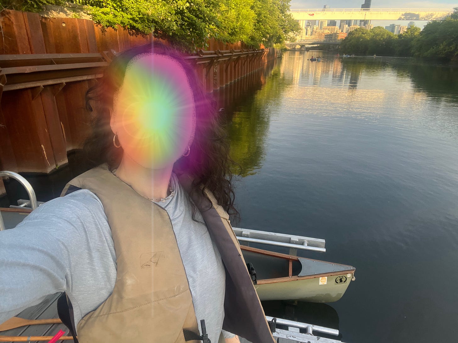 Elena is taking a selfie during the afternoon on a dock on the Chicago River. She is preparing to go canoeing; the boat is in the background. She is wearing a long-sleeved blue sun shirt and a beige life jacket. She has gold hoops in her ears, and her hair is down and wavy. Her face is blurred out with a rainbow lens flare.
