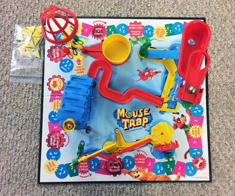 May include: A Mouse Trap board game with a blue, yellow, and red mouse trap. The game board has a yellow and red cat, a green frog, and a red mouse. The game board has the words 'Mouse Trap' printed on it. The game board is on a light brown surface.