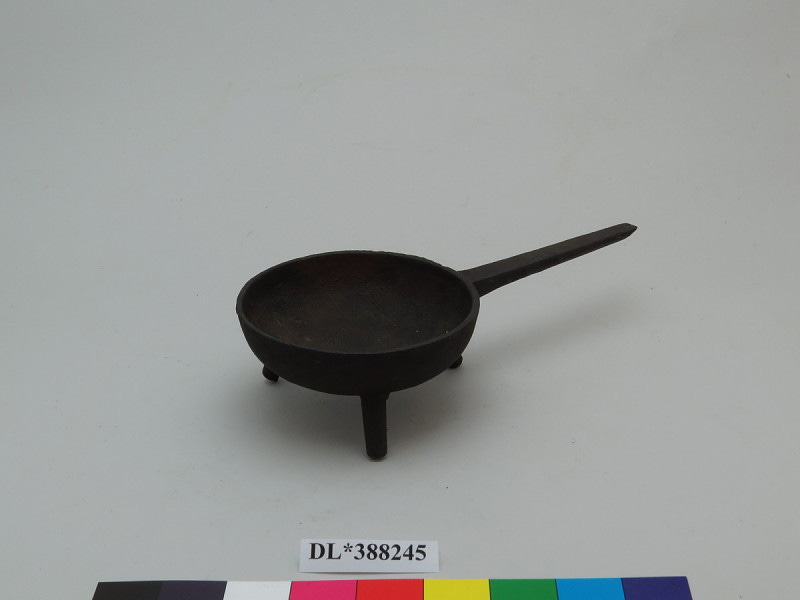 Miniature cast iron skillet on three legs. Circular bowl with rounded bottom, sitting on three tapered legs. Thin, tapered handle on one side, flattened on top. No mark.