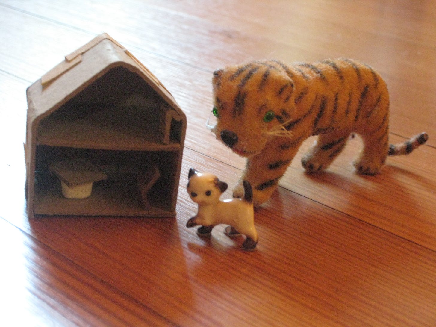 A small handcrafted dollhouse made of cardboard, a stuffed tiger, and a porcelain kitten figurine, positioned on a wooden floor