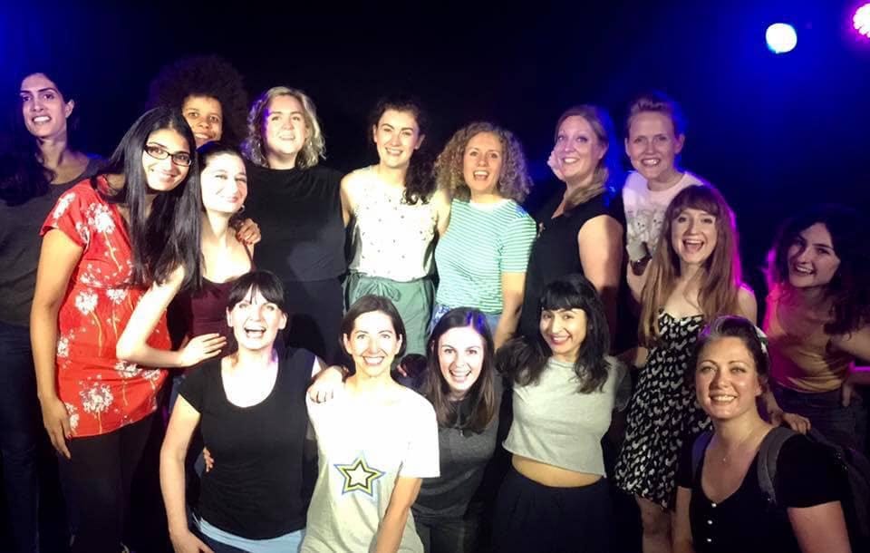A big group of women stand and kneel on stage smiling at the camera