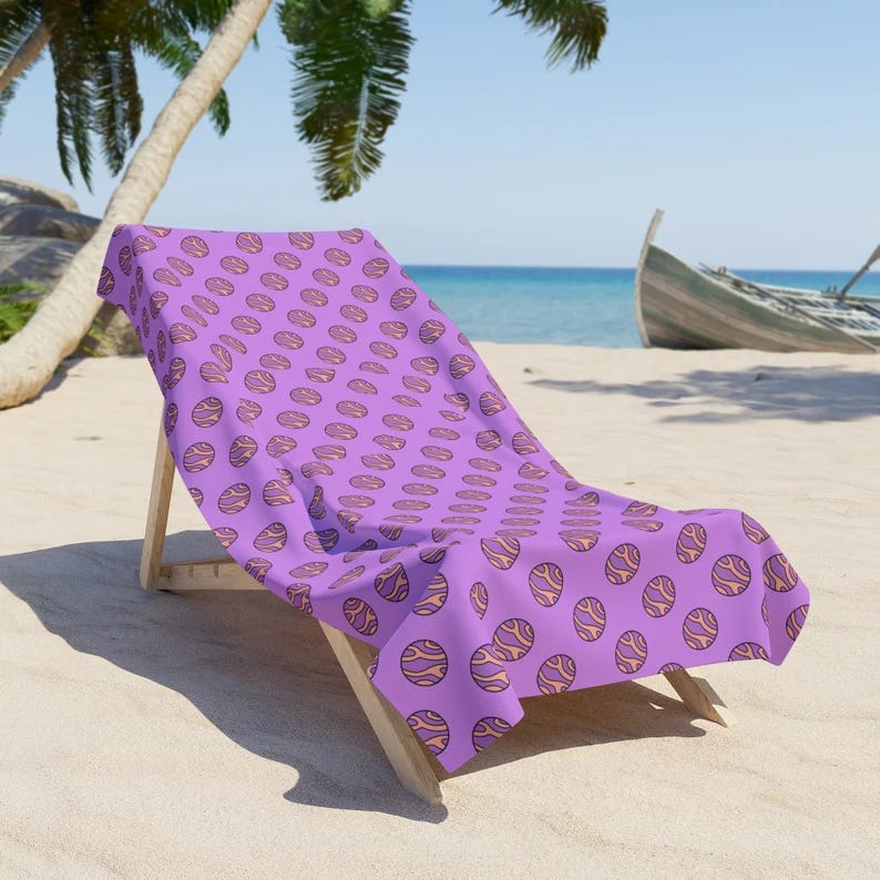 A purple beach towel with Planet Nude logos all over it draped over a beach chair in a serene tropical beach setting. 