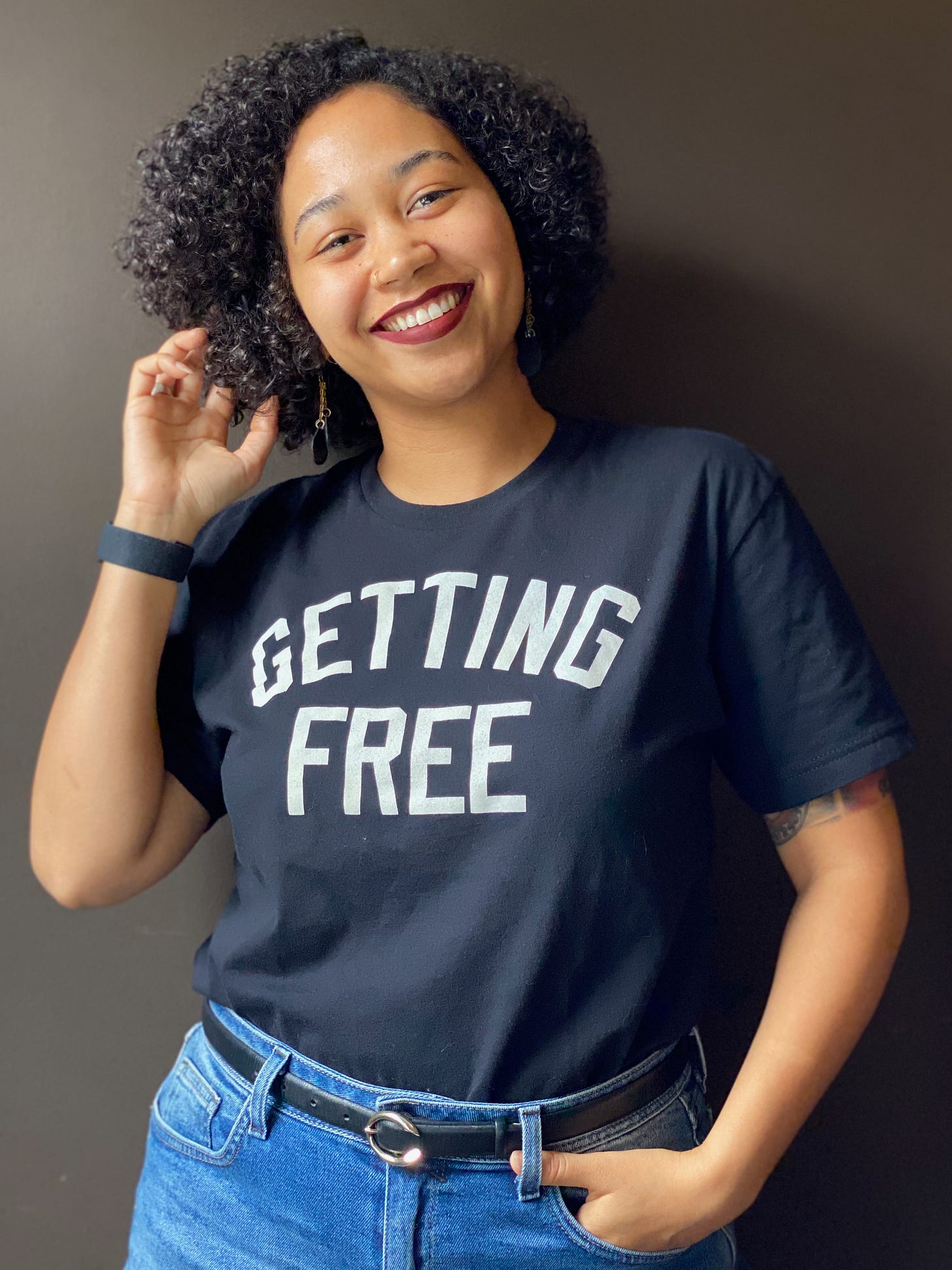 Anela stands against a wall smiling, with a red lipstick on touching her big curly hair, wearing a tee shirt that reads "getting free"