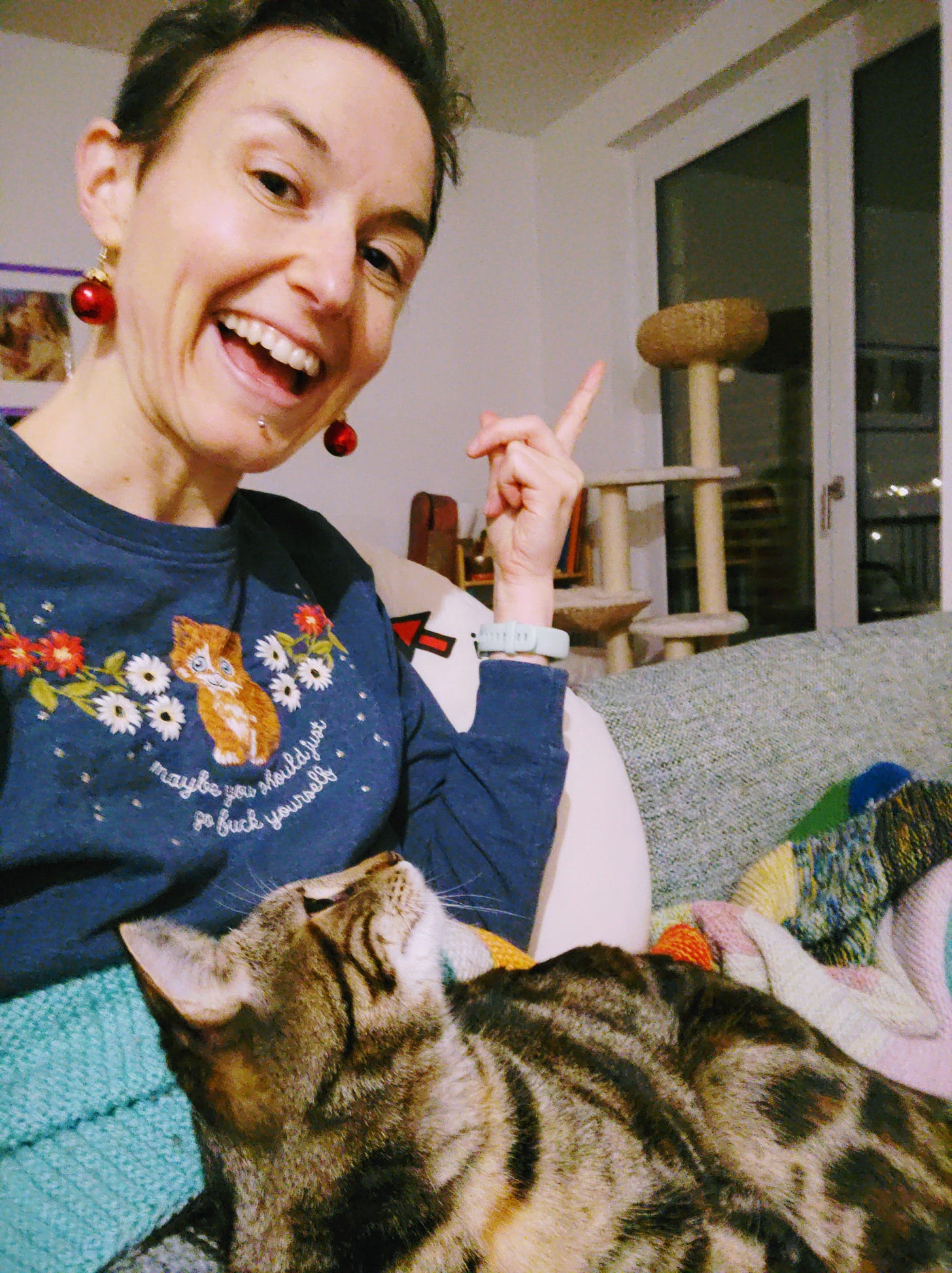On the left is a white woman with short brown hair and red earrings that look like small Christmas tree ornaments. She is smiling and pointing at a scratching post behind her that is visible in the picture. She is sitting on the couch with her cat, Luna, who is looking up at the woman.