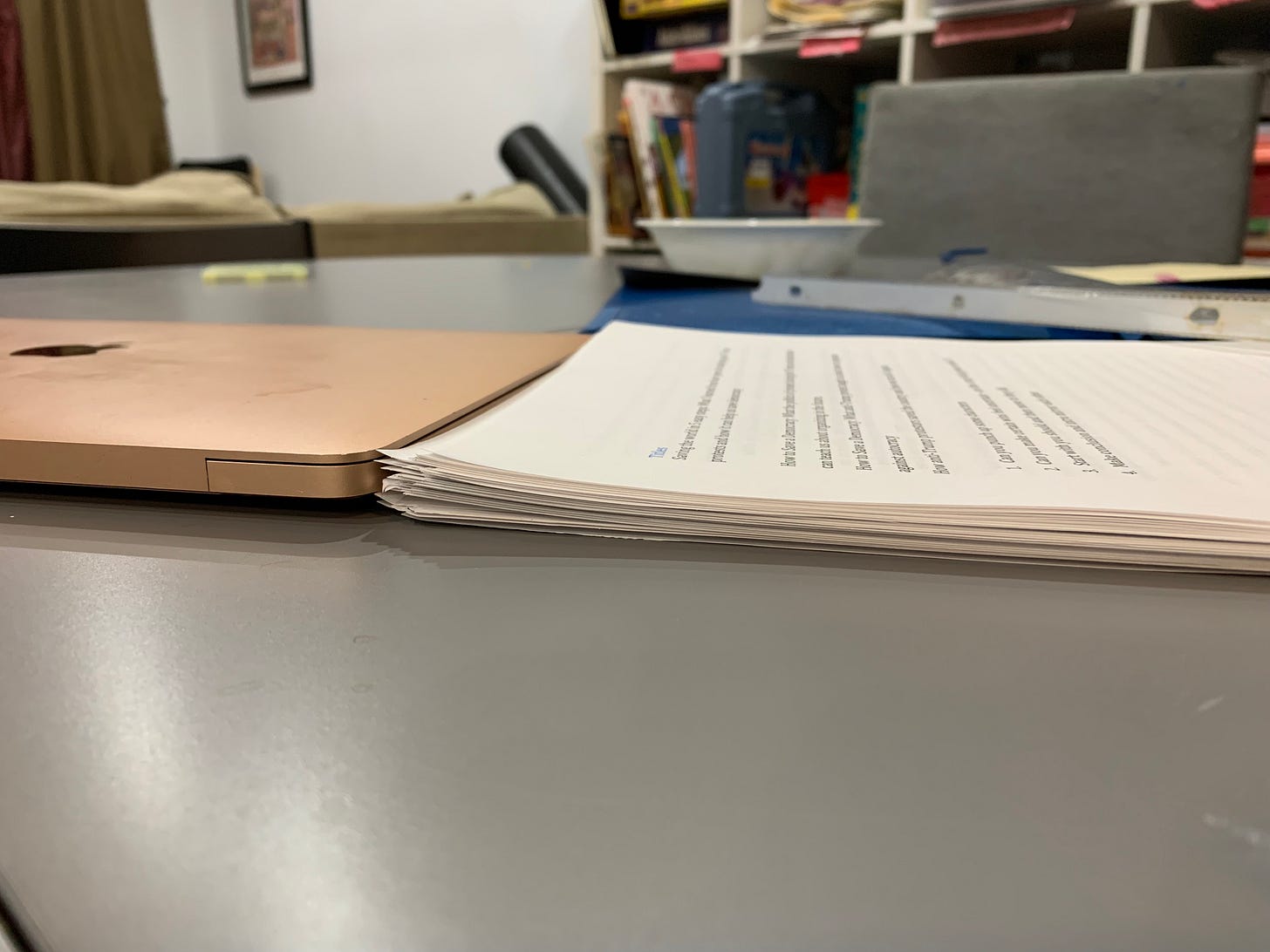 A stack of papers sitting next to a laptop on a table with a couch and bookshelf in the background.