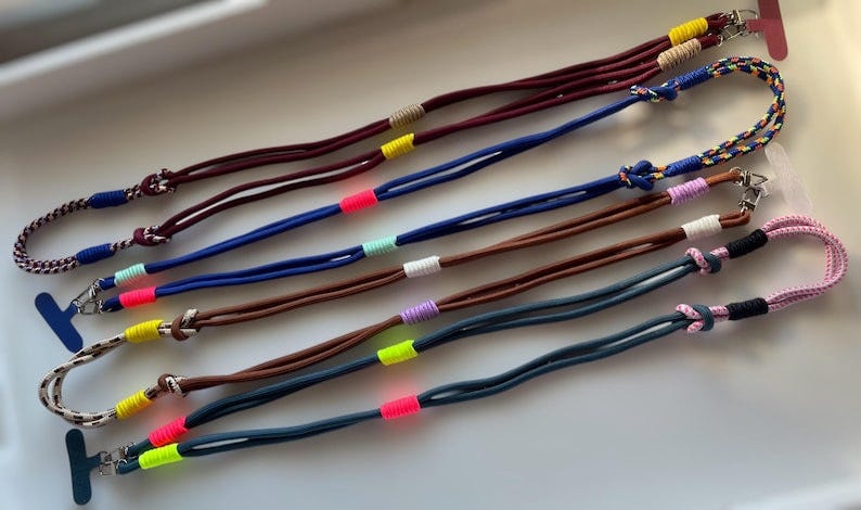 May include: Five pairs of colorful lanyard necklaces.  Each necklace features two cords in a dark color (maroon, navy, brown, dark teal) accented with brightly colored neon threads (yellow, pink, light green, purple).  The cords are knotted at the ends and feature a small metal clasp. These are perfect for holding keys, ID badges, or other small items.