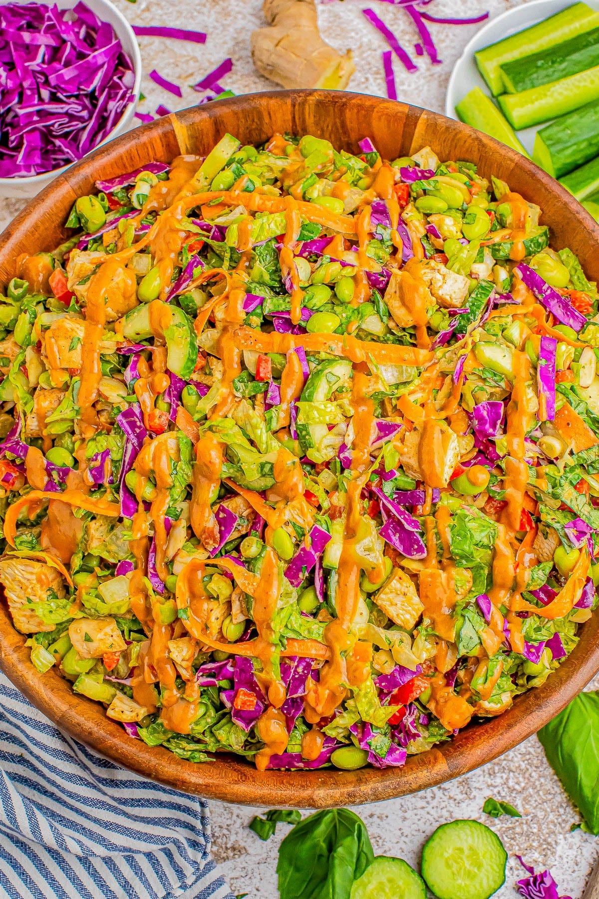 A wooden bowl filled with a colorful vegetable salad topped with a drizzle of orange dressing. Sliced cucumbers, chopped red cabbage, greens, and ginger surround the bowl. A wooden bowl filled with a colorful vegetable salad topped with a drizzle of orange dressing. Sliced cucumbers, chopped red cabbage, greens, and ginger surround the bowl.