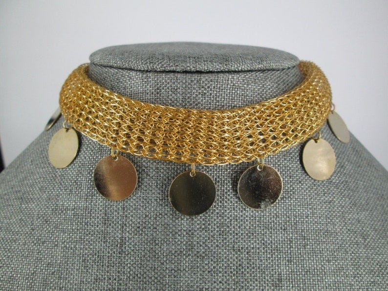 May include: A gold-toned choker necklace with a woven mesh design. The necklace features several dangling, circular gold-colored charms. The jewelry is displayed on a gray mannequin head.