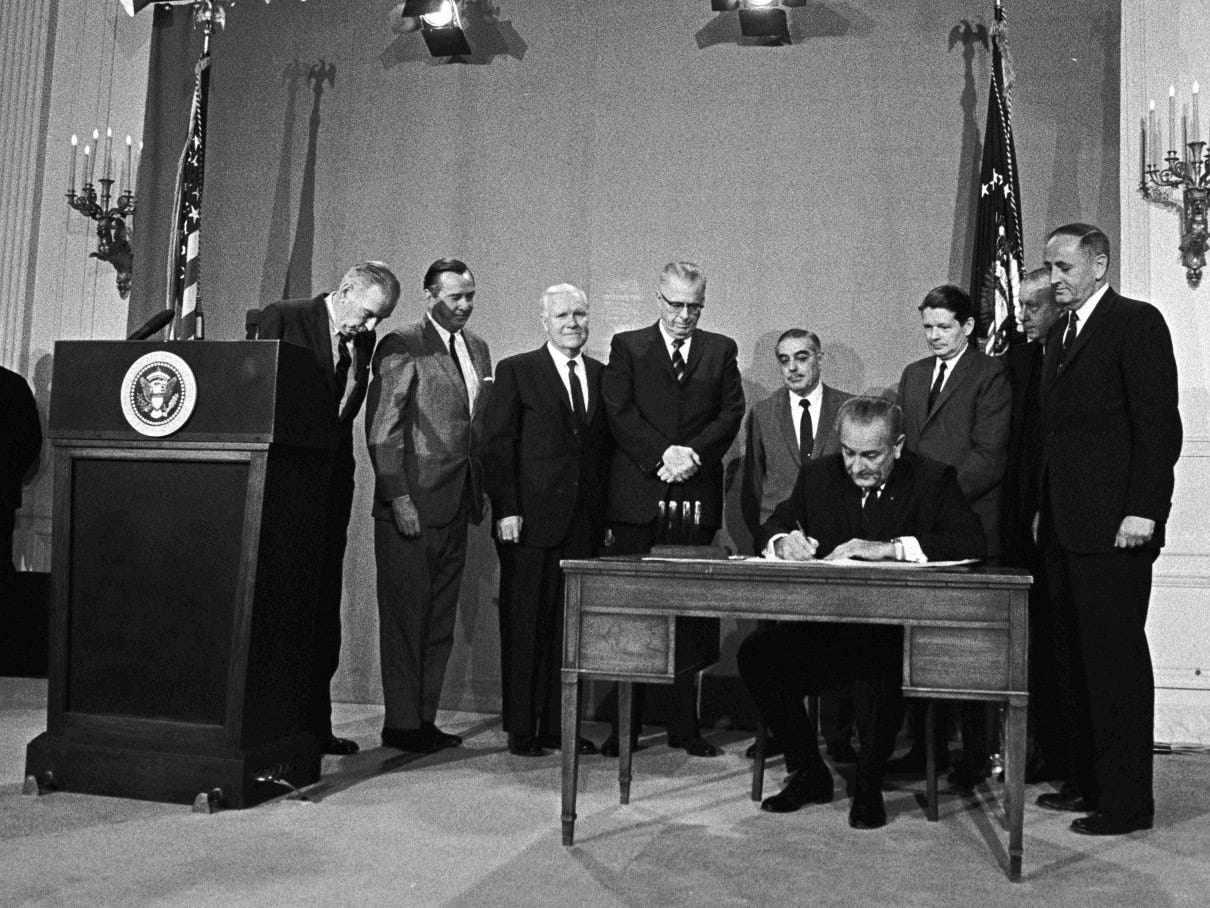 President Lyndon B. Johnson is seated at a desk with eight men in suits standing behind him. Johnson is in the act of signing a bill into law. On the left of the photo is a podium with the presidential seal emblazoned on the front, and in the background are two American flags flanking the scene.