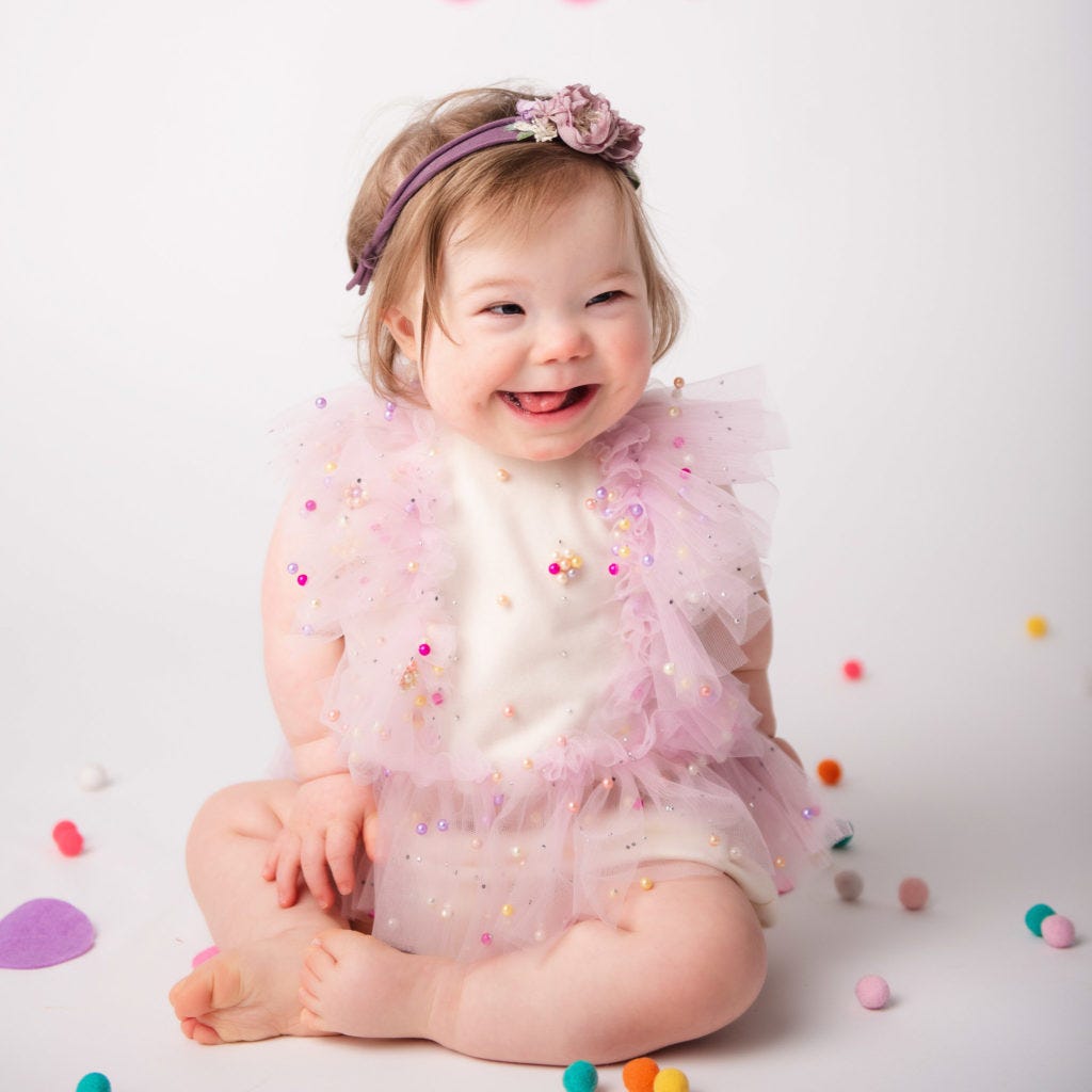 A baby girl sits crosslegged on white studio smiling in white pink dress with a purple headband.