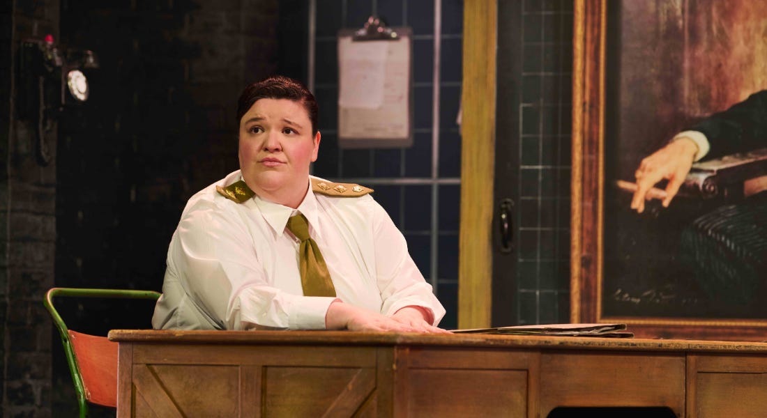 A woman wearing a shirt and gold tie sat at a desk on stage A woman wearing a shirt and gold tie sat at a desk on stage