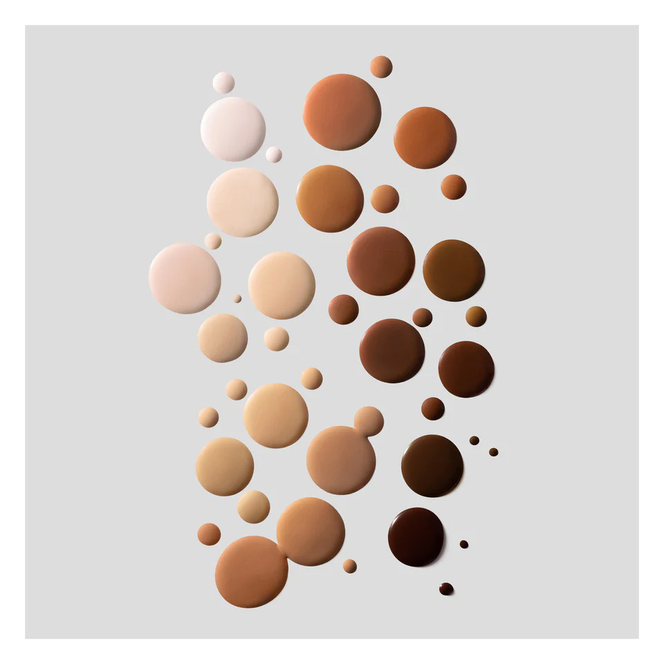 Artistically arranged droplets of Victoria Beckham Beauty Foundation Drops in a wide range of shades, from fair to deep espresso, displayed on a light gray background.