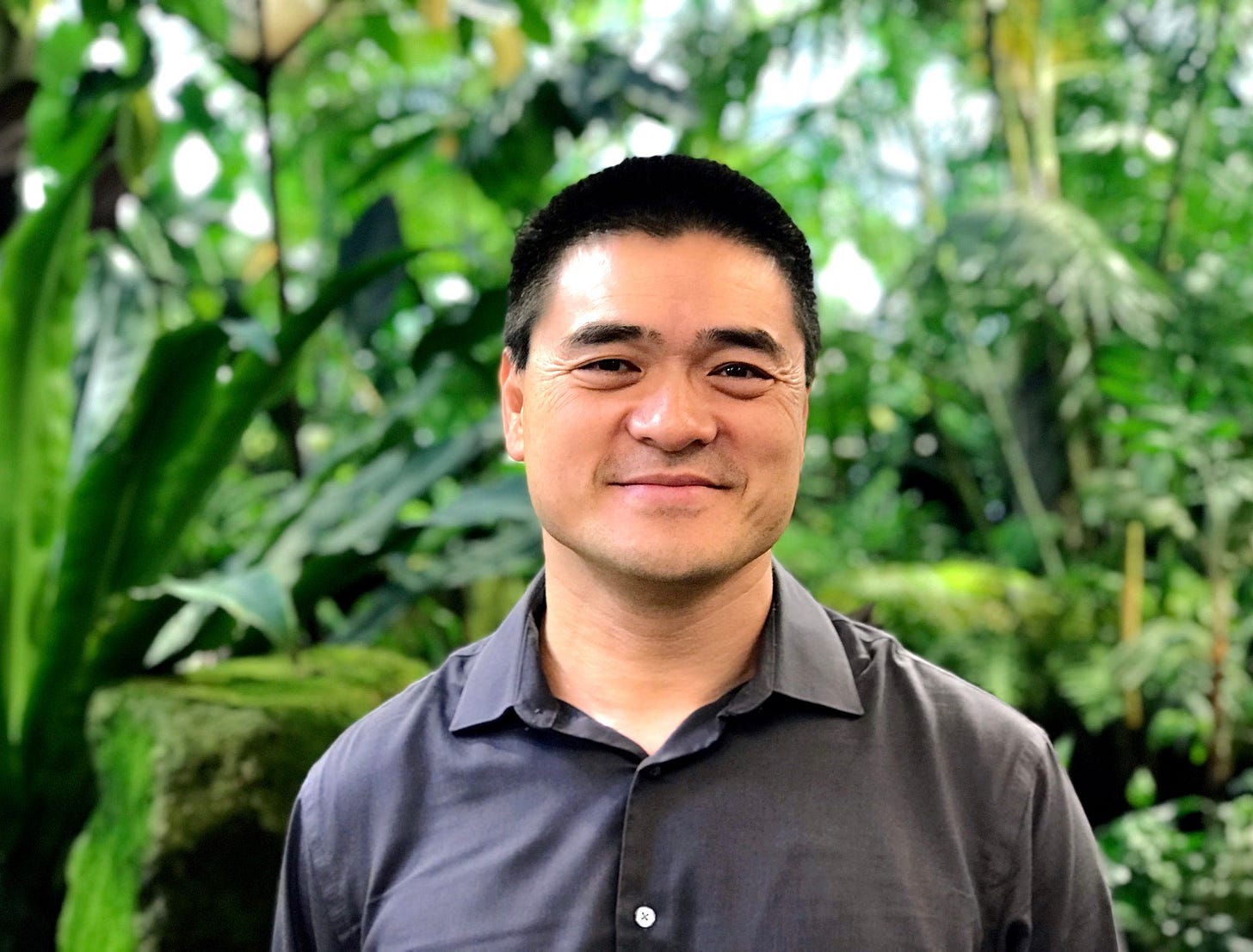 A headshot of Rich Hua. He is smiling and wearing a dark blue/gray shirt. Behind him are green plants and trees.