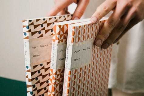 A selection of diaries and planners. Some are stacked up like in a bookshelf with labels on them, a woman sits and reads another, another is bright olive green.