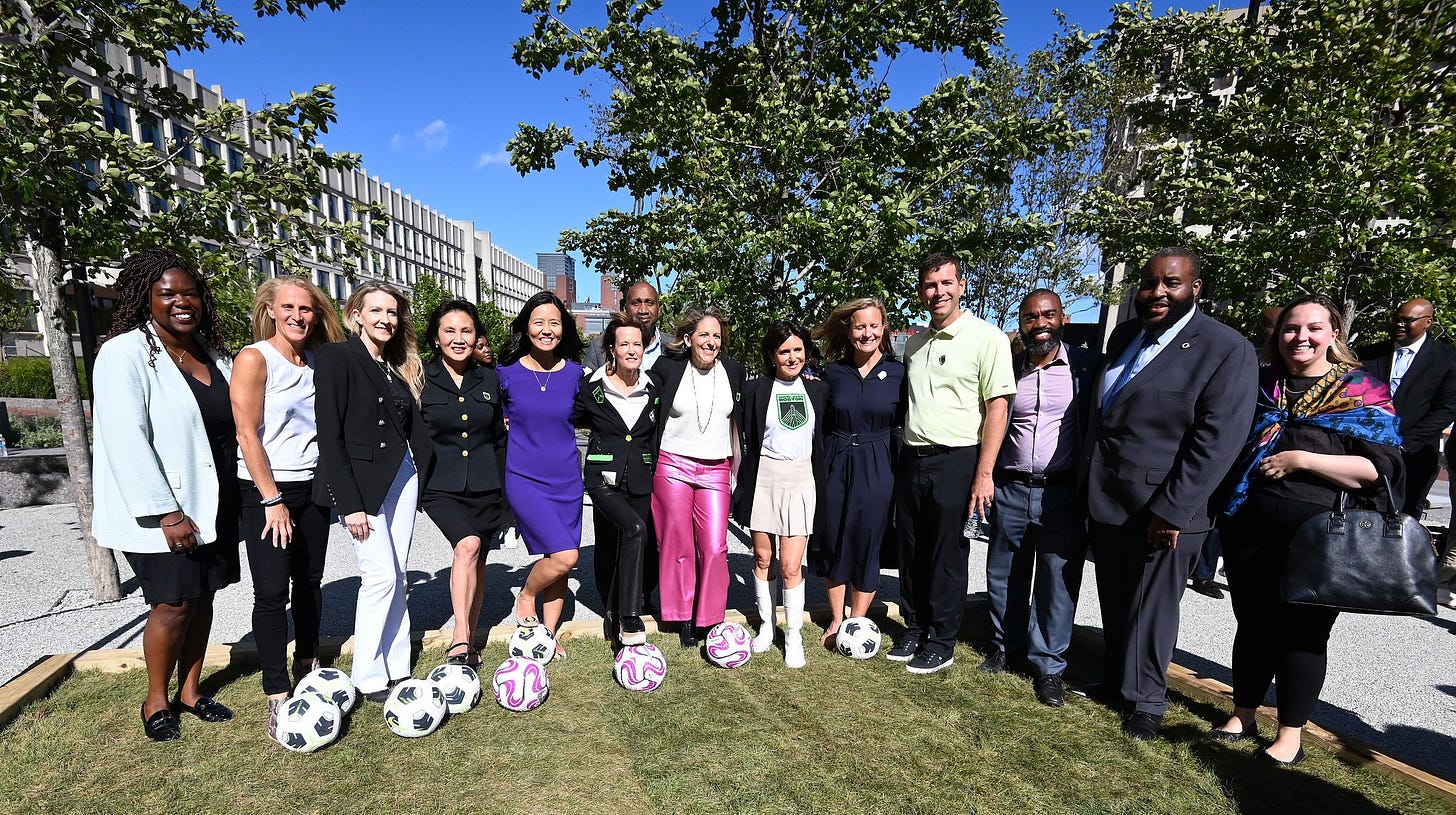About a dozen people pose for a picture on a miniature grass field, with soccer balls on the ground in front of the group