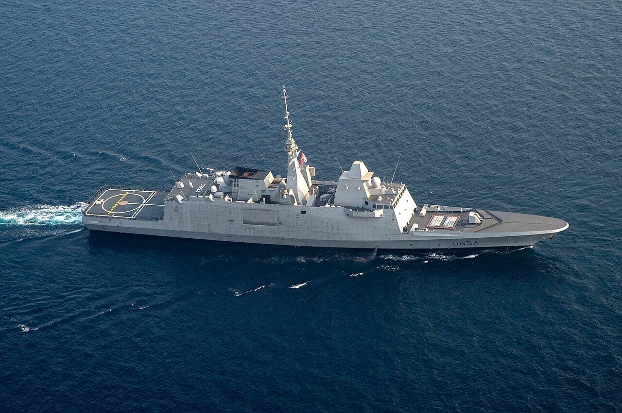 Aerial view of a grey French frigate with hull number D654 underway on calm blue water, a helicopter deck at its stern. Aerial view of a grey French frigate with hull number D654 underway on calm blue water, a helicopter deck at its stern.