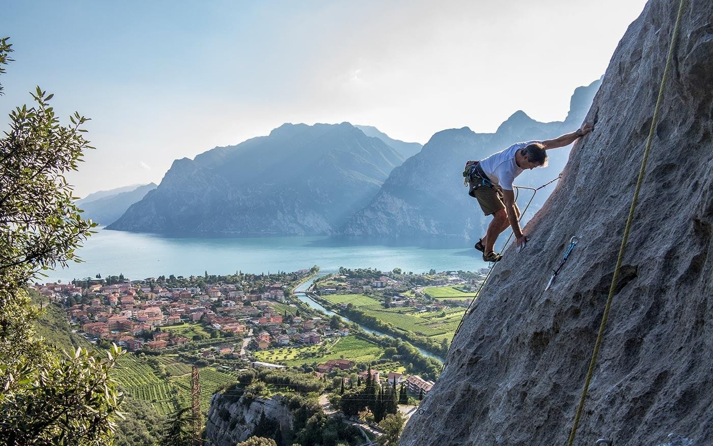 A climber ascending a rock wall near Arco, Italy, with the mountains surrounding Lake Garda visible in the distance. A climber ascending a rock wall near Arco, Italy, with the mountains surrounding Lake Garda visible in the distance.