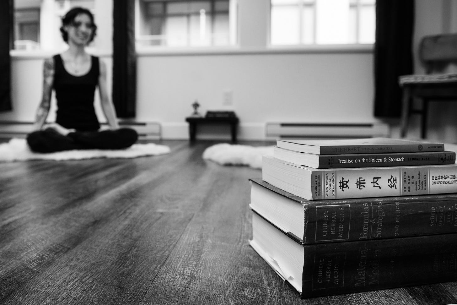 A stack of Chinese Medicine books in the foreground, with me sitting in lotus posture on the floor in the background. Black-and-white photo.