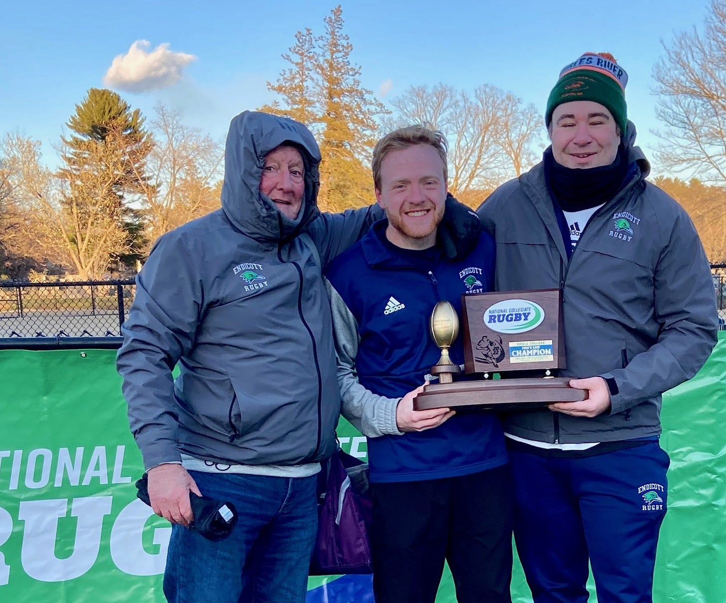 A celebratory photo of Ryan with his former rugby coaches, Desmond (far right) and Charles (far left), at a rugby championship event. They are standing outdoors on a chilly day, dressed in Endicott Rugby jackets and winter gear. Ryan, positioned in the center, is holding a championship trophy with Desmond. The trophy features a gold rugby ball and the words "National Collegiate Rugby Champion." The group is smiling, showing pride in their achievement, with a green tournament banner visible in the background.
