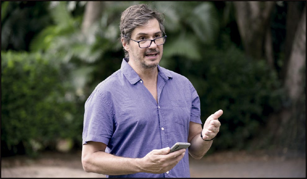 Homem adulto em ambiente externo, com vegetação ao fundo. Ele usa camisa de manga curta azul e óculos apoiados no rosto, levemente abaixo da linha dos olhos. Segura um celular em uma das mãos e gesticula com a outra, como se estivesse explicando algo ou conversando. A expressão é atenta e envolvida, e o cenário sugere um parque ou jardim.